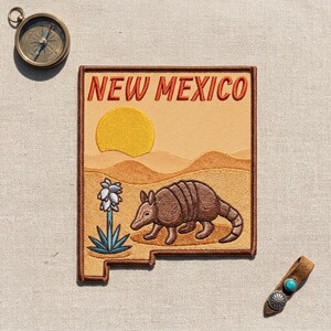 May include: A rectangular embroidered patch shaped like the state of New Mexico, featuring a brown armadillo, a yellow sun, desert dunes, and a blue yucca plant. The text NEW MEXICO is stitched in red at the top. Includes a compass and leather tab.