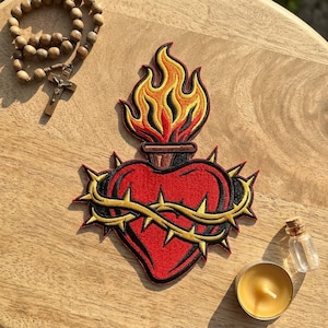 May include: An embroidered patch featuring a red heart encircled by a gold crown of thorns, topped with a stylized flame in yellow, orange, and red. The patch is displayed alongside a wooden rosary with a crucifix and a small glass vial.