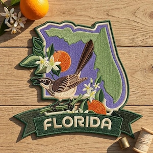 May include: An embroidered patch shaped like the state of Florida with a purple background, featuring a brown mockingbird perched on a branch with white orange blossoms and orange fruit. A green banner at the bottom displays the text FLORIDA.
