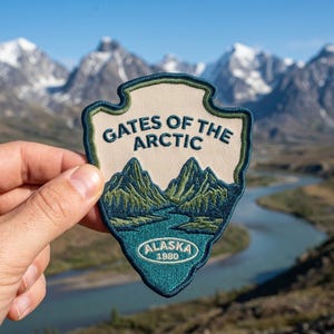 May include: A shield-shaped embroidered patch featuring a cream-colored background with dark blue text reading GATES OF THE ARCTIC. Below the text is a landscape illustration of green mountains and a blue river, with an oval containing ALASKA 1980.
