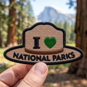 May include: An embroidered patch shaped like a ranger hat in tan with a brown brim. It features the black text I, a green heart symbol, and the text NATIONAL PARKS in black. The entire patch is outlined with a thick black stitched border.