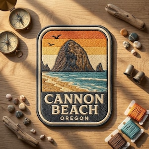 May include: A rectangular embroidered patch featuring a landscape of Haystack Rock at Cannon Beach, Oregon. The design includes a sunset sky in orange and yellow, a blue ocean, and sandy shores. The text reads CANNON BEACH OREGON in cream letters.