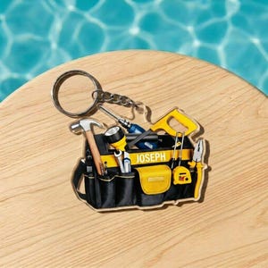 May include: A novelty keychain featuring a toolbox design with various tools, including a hammer, saw, and screwdriver. The keychain is black and yellow with the name "JOSEPH" printed on it. The keychain is attached to a silver keyring.