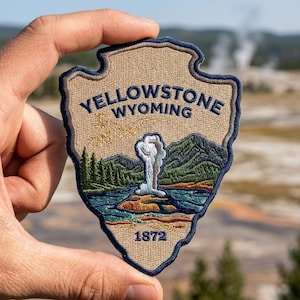 May include: A shield-shaped embroidered patch featuring a tan background with a dark blue border. The text reads YELLOWSTONE WYOMING and 1872. The design depicts a geyser erupting in a landscape with green pine trees, mountains, and blue water.