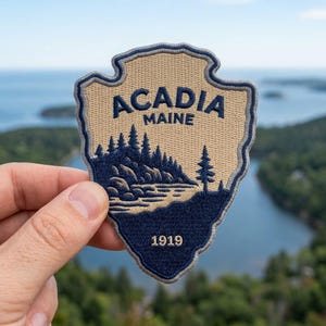 May include: A shield-shaped embroidered patch featuring a tan background with dark blue stitching. The text ACADIA MAINE is at the top, with a landscape of pine trees and a rocky shoreline below. The year 1919 is stitched in dark blue at the bottom.