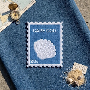 May include: A rectangular embroidered patch featuring a blue background with a white scalloped border, a white seashell illustration, and the white text "CAPE COD" above and "20¢" below, placed on a blue denim fabric surface.