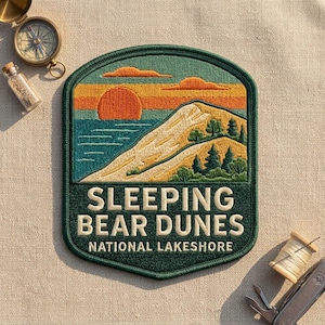 May include: A shield-shaped embroidered patch featuring a landscape of Sleeping Bear Dunes National Lakeshore with a sunset, sand dunes, and pine trees. The text reads SLEEPING BEAR DUNES NATIONAL LAKESHORE in cream-colored capital letters.