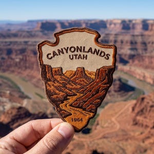 May include: A shield-shaped embroidered patch featuring a landscape of Canyonlands, Utah. The patch displays the text CANYONLANDS UTAH at the top and 1964 at the bottom, with brown and tan thread depicting a winding river and canyon rock formations.