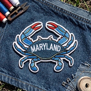 May include: An embroidered patch featuring a blue crab with red-tipped claws and the word MARYLAND stitched in white across its shell. The patch has a white border and is placed on dark blue denim fabric near sewing supplies.