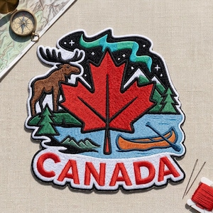 May include: A decorative embroidered patch featuring a large red maple leaf, a brown moose, a green forest, a blue lake with an orange canoe, and a teal aurora borealis against a black starry sky, with the word CANADA in red letters at the bottom.