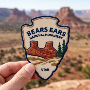 May include: An embroidered patch shaped like an arrowhead with a cream background and dark blue border. It features the text BEARS EARS NATIONAL MONUMENT and UTAH, with a graphic of red rock formations and green pine trees in the center.