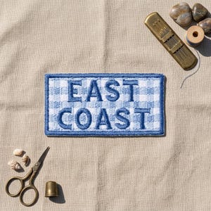 May include: A rectangular embroidered patch featuring a blue and white gingham pattern with the text EAST COAST in bold blue letters. The patch is surrounded by sewing tools including a brass needle case, a thimble, scissors, and small seashells.
