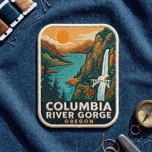 May include: A rectangular embroidered patch featuring a landscape of the Columbia River Gorge in Oregon. The design shows a waterfall, a bridge, mountains, and a sun in shades of green, blue, and orange. The text reads COLUMBIA RIVER GORGE OREGON.