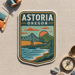 May include: A rectangular embroidered patch featuring the text ASTORIA OREGON above a landscape scene. The design depicts a bridge spanning a body of water with green hills, a setting sun, and orange clouds, all bordered by a thick yellow and cream frame.