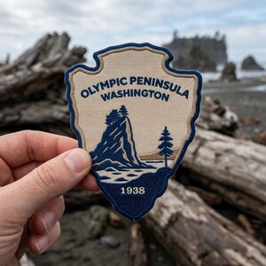 May include: A shield-shaped embroidered patch featuring a beige background with a dark blue border. The text reads OLYMPIC PENINSULA WASHINGTON and 1938. The design depicts a dark blue coastal rock formation with trees and a single pine tree.
