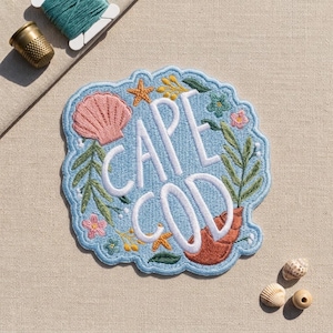 May include: An embroidered patch featuring the text CAPE COD in white letters, surrounded by a light blue border. The design includes a pink seashell, a brown conch shell, orange starfish, green leafy branches, and small pink and yellow flowers.