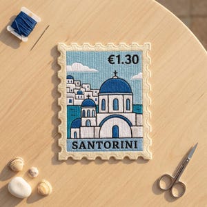 May include: A rectangular embroidered patch shaped like a postage stamp featuring a blue-domed church in Santorini, Greece. The design includes white buildings, a blue sea, and the text "€1.30" and "SANTORINI" stitched in black thread.