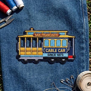 May include: An embroidered patch of a San Francisco cable car featuring yellow, blue, and black thread. The text reads SAN FRANCISCO CABLE CAR SINCE 1873. The patch is placed on blue denim fabric near sewing supplies including thread and scissors.