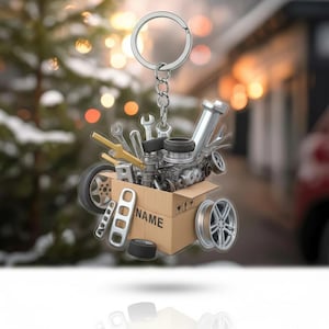 May include: A silver keychain featuring a miniature cardboard box filled with car parts and tools. The box has the word "NAME" printed on it. The keychain includes a car engine, wrenches, and tires. The background is blurred.
