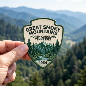 May include: A shield-shaped embroidered patch featuring the text GREAT SMOKY MOUNTAINS NORTH CAROLINA TENNESSEE and 1934. The design depicts a green mountain range, a winding river, and evergreen trees against a cream-colored background.
