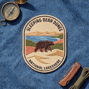 May include: An oval embroidered patch featuring a brown bear walking across a landscape of tan sand dunes, green hills, and a blue lake. The text "SLEEPING BEAR DUNES" is arched at the top and "NATIONAL LAKESHORE" is arched at the bottom in black.