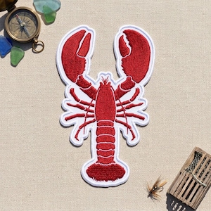 May include: A red embroidered lobster patch with a white border. The patch features detailed stitching of the lobster's claws, legs, and segmented tail. It is displayed alongside a brass compass, sea glass pieces, and a small wooden trap.