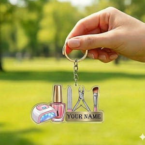 May include: A clear acrylic keychain featuring nail care tools: a nail polish bottle, a UV lamp, a nail file, clippers, and a makeup brush. The keychain has a gold-colored ring and chain. The text "YOUR NAME" is printed on a gold-colored rectangle.