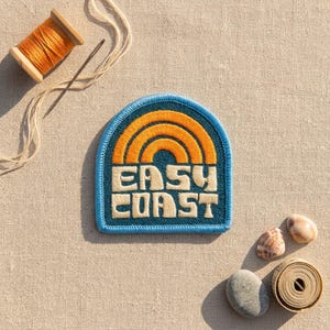 May include: An embroidered patch with a blue background, an orange rainbow graphic, and the text "EASY COAST" in cream. Included are a wooden spool of orange thread, a metal needle, two seashells, and a coiled measuring tape on a beige fabric.