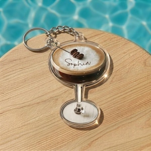 May include: Clear acrylic keychain shaped like a cocktail glass, featuring a coffee drink with foam and coffee beans. The name "Sophia" is written on the foam. The keychain has a silver chain and ring, set on a wooden surface.