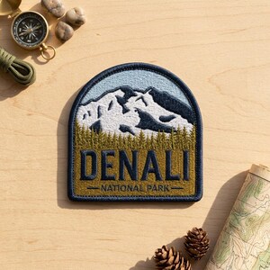 May include: An embroidered patch with a dark blue border featuring a landscape of snow-capped mountains against a light blue sky, a forest of green pine trees, and the text DENALI NATIONAL PARK in dark blue letters on a mustard yellow background.