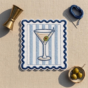 May include: A square embroidered patch with a scalloped dark blue border featuring a martini glass with a green olive on a toothpick, centered on a light blue and white vertical striped background, accompanied by a brass jigger and blue thread.