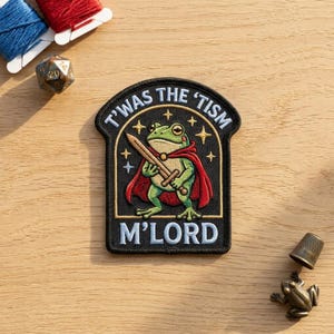 May include: An embroidered patch featuring a green frog wearing a red cape and holding a sword. The black background includes gold stars and the white text T'WAS THE 'TISM M'LORD. The patch has a rounded top and a rectangular base.