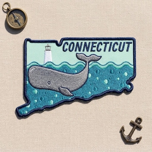 May include: An embroidered patch shaped like the state of Connecticut featuring a gray whale swimming in dark teal water with bubbles, a white lighthouse in the background, and the text CONNECTICUT in dark blue letters on a light teal sky.