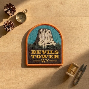 May include: An embroidered patch featuring a depiction of Devils Tower in Wyoming. The design includes a light-colored rock formation against a blue sky, a dark green forest, and the yellow text "DEVILS TOWER WY" on a dark brown background.