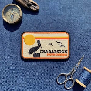 May include: A rectangular embroidered patch featuring a black pelican silhouette, a yellow sun, and three flying birds against a cream background with orange stripes. The text reads CHARLESTON SOUTH CAROLINA in black and orange lettering.