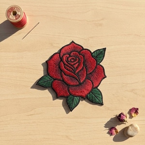 May include: An embroidered rose patch featuring vibrant red petals with dark outlines and green leaves. The patch is accompanied by a wooden spool of red silk thread, a sewing needle, two dried rose petals, and a small smooth stone.