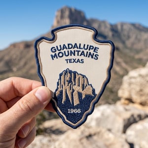 May include: A shield-shaped embroidered patch featuring a cream-colored background with a dark blue border. The text reads GUADALUPE MOUNTAINS TEXAS in dark blue, with a stylized mountain graphic and the year 1966 embroidered in white at the base.