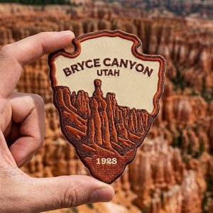 May include: A shield-shaped embroidered patch featuring a cream-colored background with brown text reading BRYCE CANYON UTAH and 1928. The lower portion displays a detailed brown stitched landscape of hoodoos and rock formations.