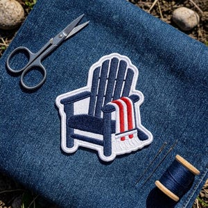 May include: An embroidered patch featuring a dark blue Adirondack chair with a white and red striped blanket draped over the arm, placed on blue denim fabric alongside silver metal embroidery scissors and a spool of dark blue thread.
