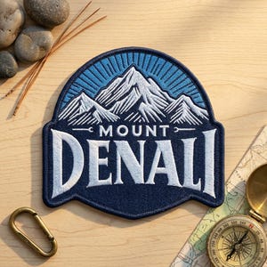 May include: A navy blue embroidered patch featuring a white mountain range graphic above the text MOUNT DENALI in bold white lettering. The design includes a light blue sunburst pattern behind the mountains and a dark blue stitched border.