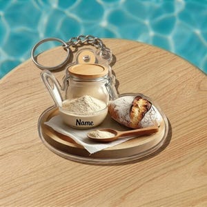 May include: A clear acrylic keychain featuring a miniature bread-making scene. It includes a small loaf of bread, a bowl of flour with the word "Name", a jar, and a wooden spoon. The keychain has a silver chain and ring.