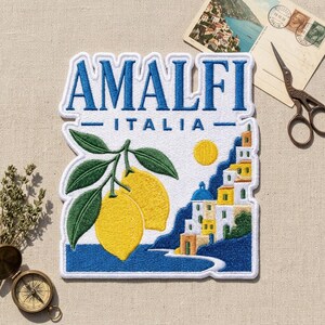 May include: A rectangular embroidered patch featuring the text AMALFI ITALIA in blue. The design includes two yellow lemons with green leaves on the left and a coastal scene with white buildings, yellow accents, and a yellow sun on the right.
