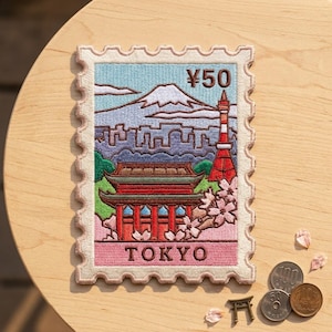May include: A rectangular embroidered patch shaped like a postage stamp featuring a scene of Tokyo. The design includes a red and white tower, a temple, cherry blossoms, and a mountain. The text ¥50 and TOKYO are stitched in brown thread.
