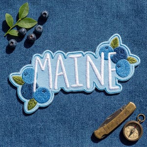 May include: An embroidered patch featuring the word MAINE in white letters on a light blue background, decorated with blue blueberry clusters and green leaves, accompanied by loose blueberries and a brass pocket knife with a compass.