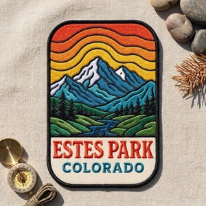 May include: A rectangular embroidered patch featuring a mountain landscape with a blue river, green hills, and a sunset sky with orange and yellow waves. The text ESTES PARK COLORADO is stitched in red and blue letters on a cream background.