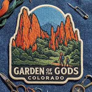 May include: An embroidered patch featuring a landscape of orange rock formations, green trees, and a blue sky. Two figures hike on a path. The text reads GARDEN OF THE GODS COLORADO. The patch has a cream-colored border and is placed on blue denim fabric.