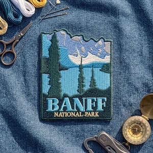 May include: An embroidered patch featuring a landscape of Banff National Park with dark green pine trees, a light blue lake, and snow-capped mountains. The text BANFF NATIONAL PARK is stitched in light blue and yellow thread on the bottom.