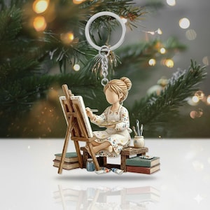 May include: A keychain featuring a figurine of an artist painting on a canvas. The artist is seated, wearing a floral apron, and holding a palette. The figurine includes a silver-colored keyring and chain. The background is blurred with Christmas tree branches and lights.