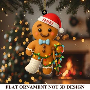 May include: A gingerbread man Christmas ornament with a red Santa hat that says "JAMES". The ornament has a blue bow tie, a wrapped leg, and a cane wrapped in Christmas lights. The text at the bottom says "FLAT ORNAMENT NOT 3D DESIGN".