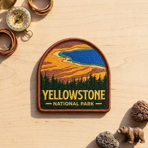 May include: An embroidered patch with a brown border featuring a landscape of Yellowstone National Park. The design shows a blue thermal pool, yellow mineral deposits, and a dark green forest. The text reads YELLOWSTONE NATIONAL PARK in yellow.